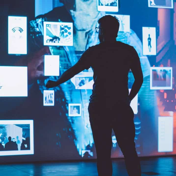 A person stands in front of a large digital screen displaying floating images and icons, interacting with the display.